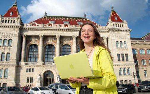 Mosolygó fiatal nő sárga laptoppal egy impozáns, klasszicista stílusú épület előtt.